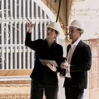 3 Personen unterhalten sich auf der Baustelle und schauen sich etwas auf einem Plan an. Jeder trägt einen Helm in unterschiedlichen Farben (weiß, grün, orange).