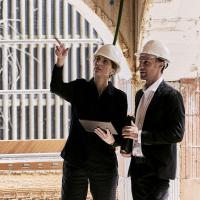 3 Personen unterhalten sich auf der Baustelle und schauen sich etwas auf einem Plan an. Jeder trägt einen Helm in unterschiedlichen Farben (weiß, grün, orange).