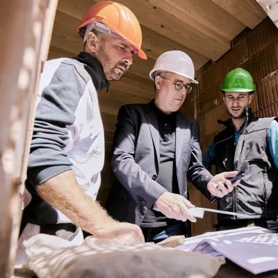 Drei Personen auf der Baustelle unterhalten sich und schauen etwas auf einem Plan an. Jeder hat einen Helm auf. Die Helme haben unterschiedliche Farben.