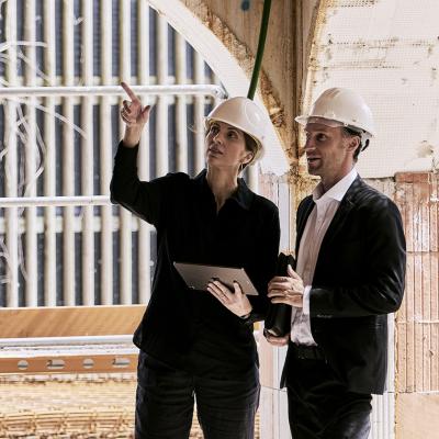 3 Personen unterhalten sich auf der Baustelle und schauen sich etwas auf einem Plan an. Jeder trägt einen Helm in unterschiedlichen Farben (weiß, grün, orange).