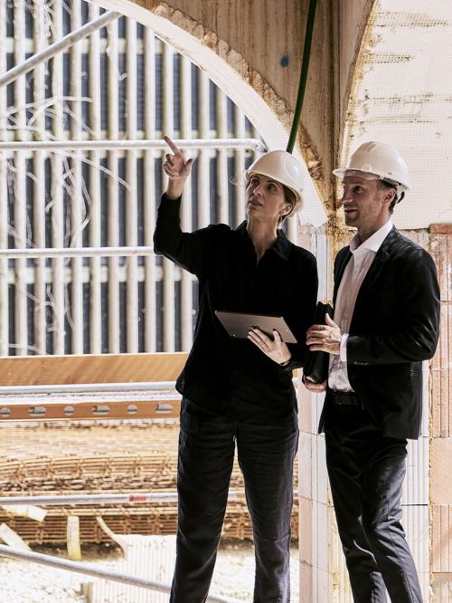 2 Personen mit weißen Helmen unterhalten sich auf der Baustelle. Sie zeigt auf etwas.