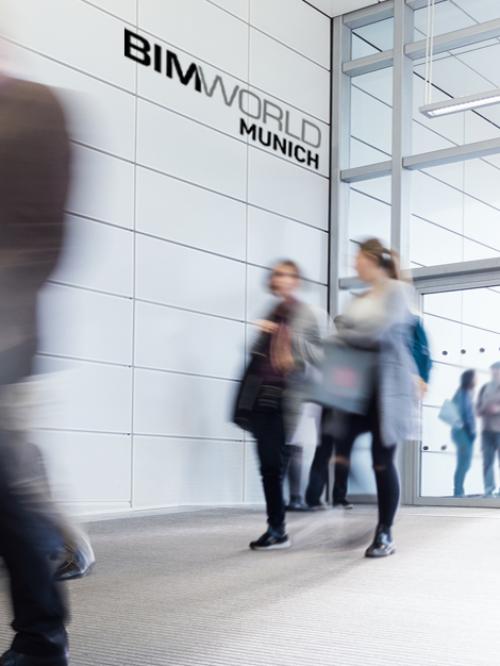 Personen  laufen vor Messegelände. Man erkennt an der Wand das Logo der BIM World Munich.
