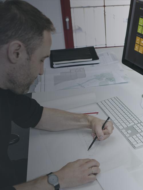 Zeichnende Person am Schreibtisch. Auf einem Bildschirm ist die Software PROJEKT PRO zu sehen.