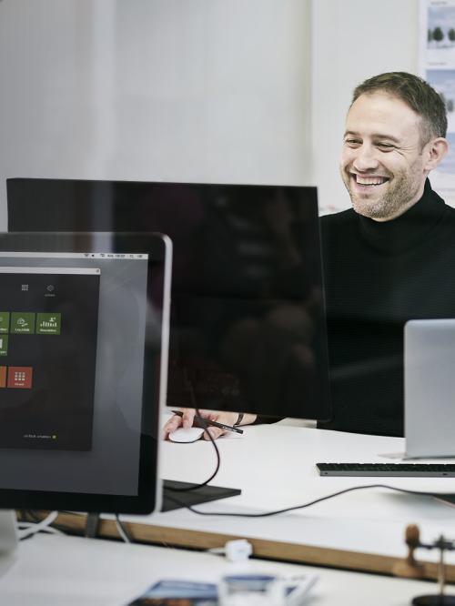 Kollegen sitzen am Schreibtisch und arbeiten mit Freude, er lacht. Man sieht die geöffnete Software PROJEKT PRO am Screen.