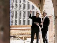 2 Personen mit weißen Helmen unterhalten sich auf der Baustelle. Sie zeigt auf etwas.