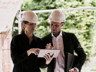 2 Personen mit weißen Helmen schauen auf der Baustelle auf ein Tablet. 