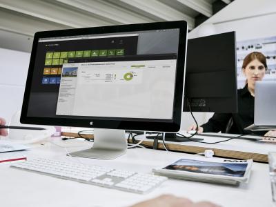 Bildschirm mit geöffneter Software von PROJEKT PRO. Im Hintergrund Architektin, die an einem anderen Schreibtisch arbeitet.